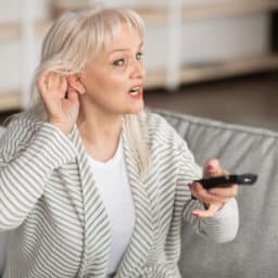 Portrait of senior woman with impaired hearing watching TV, trying to listen holding her hand near ear to hear better, sitting on couch in living room.